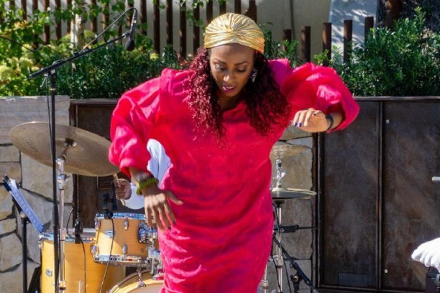 On a terrace, a Black woman, in a golden head wrap and pink dress, dances.