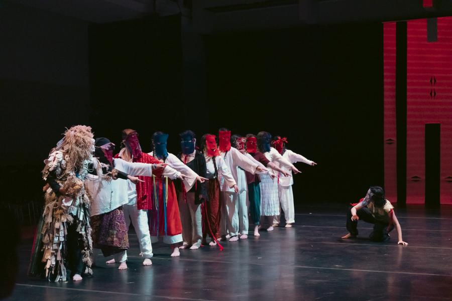 A group of masked performers stand in a line. All of them point at someone crouching on the ground in front of them.