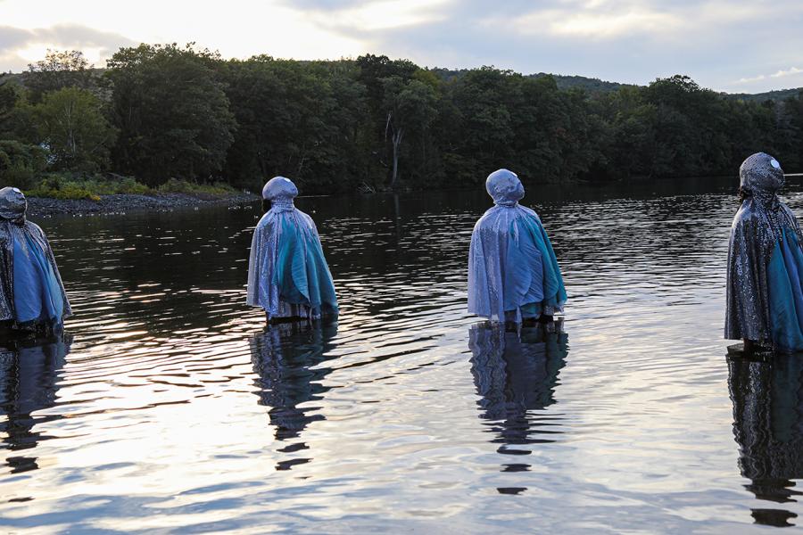 Four people, in bejeweled costumes that cover their faces, stand in water surrounded by hills.