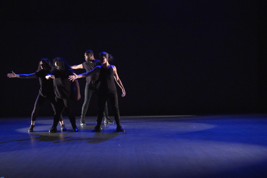 On a dim stage, four dancers, in all black, hold their arms out.
