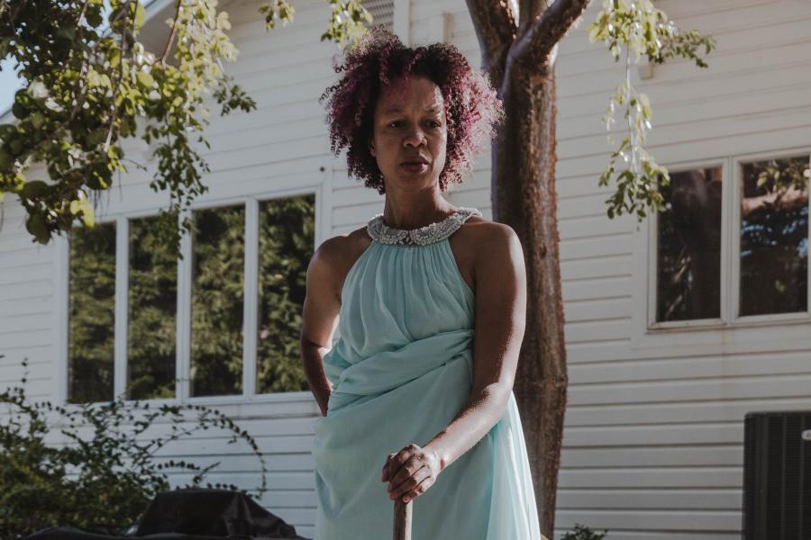A black woman stands in her yard with a broom.