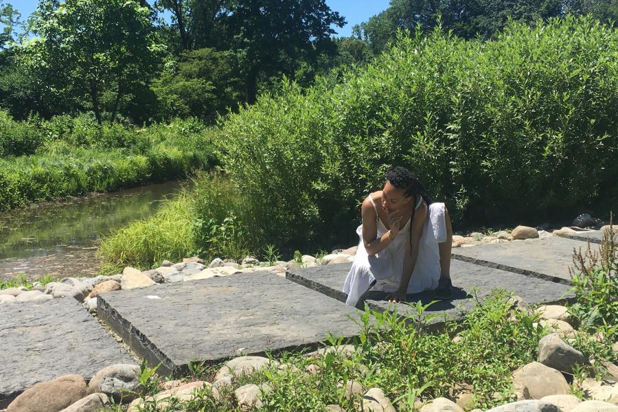 Along a stone path, a woman of color, dressed in white, crouches on one knee.