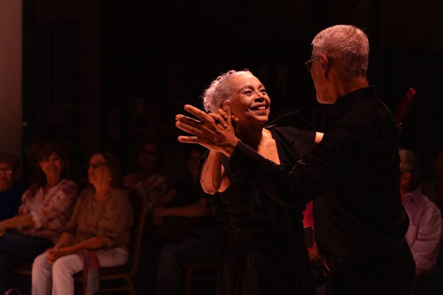An elderly couple dances, with their hands clasped. An audience watches behind them.