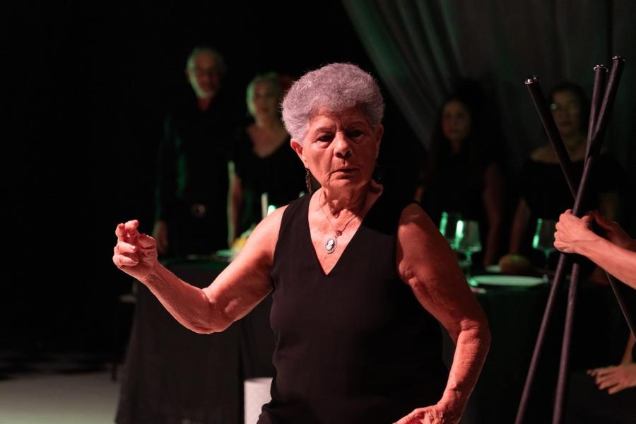 In stage lighting, an elderly woman holds a glass.