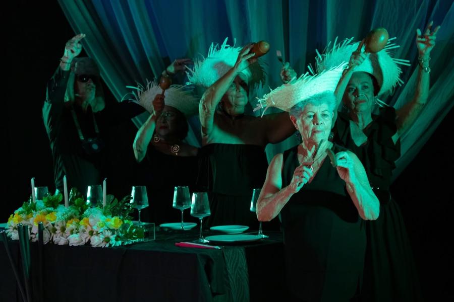 In green lighting, an elderly woman sits a table in a straw hat. More folks, in matching hats, stand behind her.