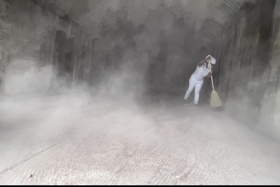 A Black woman, in white, uses a white broom to brush white dust that is moving around the room like a cloud.