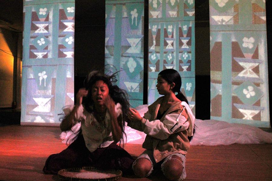 In front of a backdrops of triangular patterns, two females crouch in the shadows. One throws her hands and her hair blows as she moves. The other crosses her arms.
