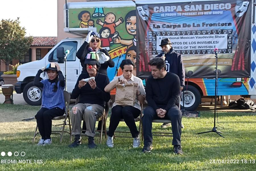 Seven bandmates together, in front of a truck with a backdrop along it. Four of them sit in folding chairs and two stand behind those four.