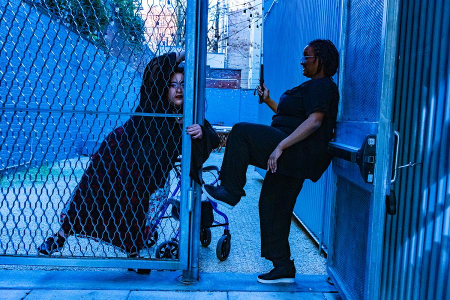 A photo of Kayla & Vanessa. On the left is Kayla Hamilton, a Black woman with locs is wearing a black shirt and pants. She is in profile and has her leg bent leaning on Vanessa’s purple walker Pluto. She is holding her folded white cane. Vanessa is a Chicana woman with caramel skin. She is leaning on a metal gate door. She is wearing a goth outfit. They are outdoors with walls in the back framing them. The photo is washed with an electric blue futuristic color.