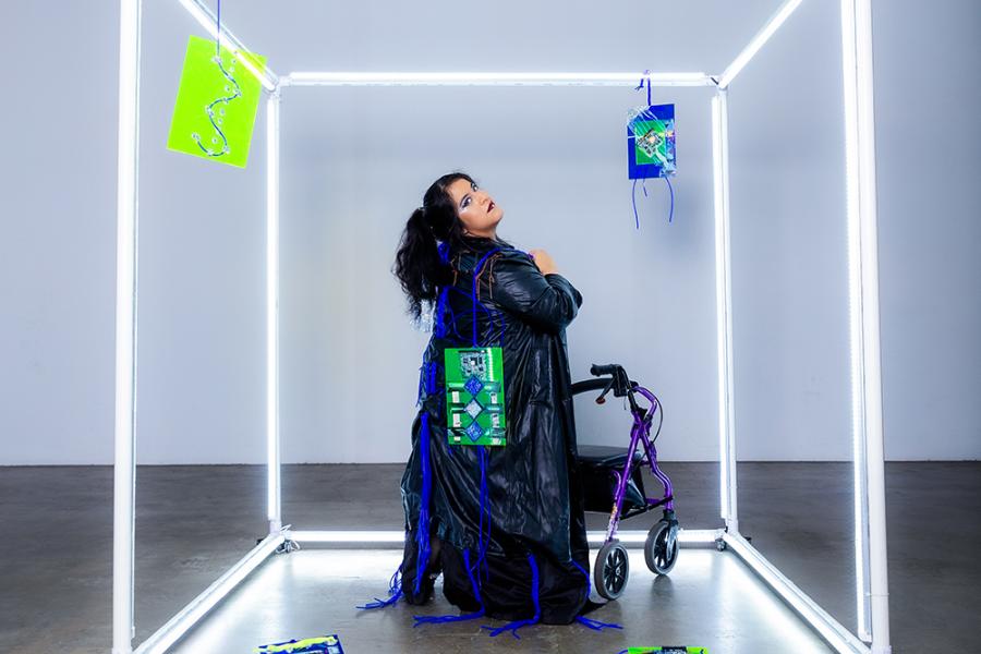 A photo of Vanessa, who is a Chicana woman with caramel skin is in an glowing cube. She is standing with her walker at a diagonal as she looks behind her as she carries one acrylic canvas behind her back. She is wearing a long sleeve shiny black holographic dress with a long leather coat.