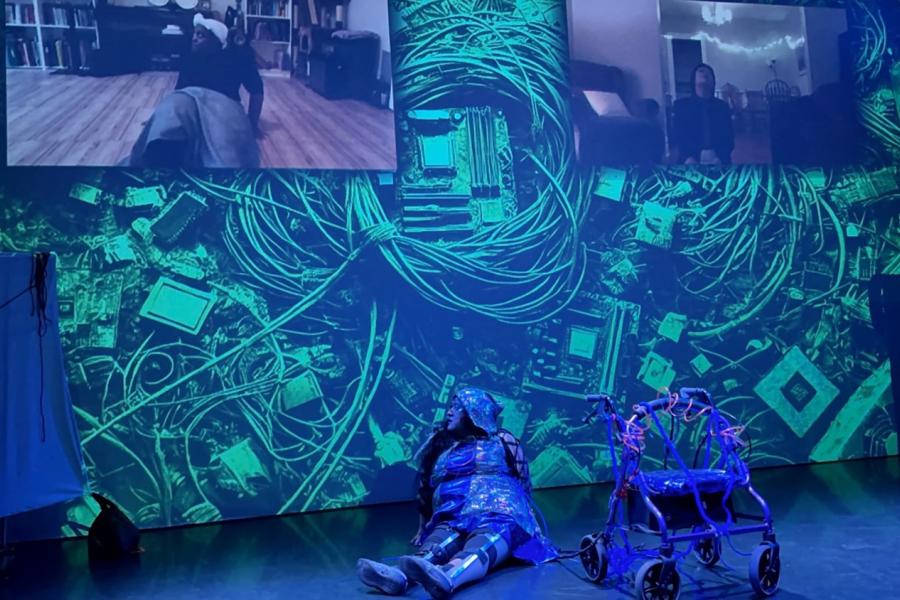 A photo of Vanessa who is a Chicana woman with caramel skin wearing a holographic blue dress hoodie leaning against a wall where there are two dancers projected above her. India who is a Black fat cisgendered woman, she is on the floor backing away. Lu who is lightskinned East Asian person is looking up from their space. The projected backdrop are wires, computer chips, cords with two pulled out blank screen on the right and left of the stage.