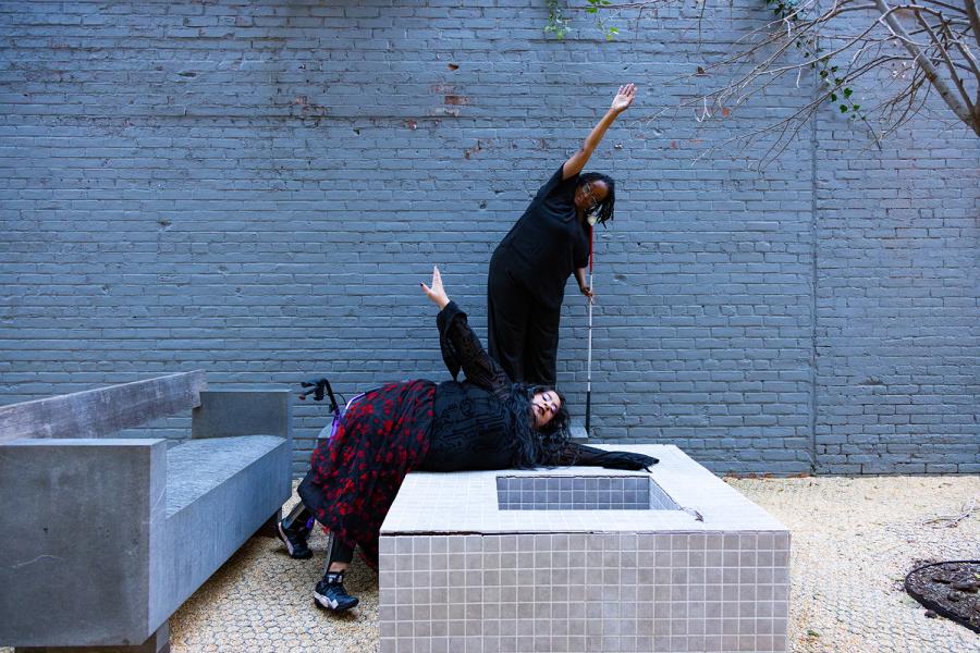 A photo of Vanessa & Kayla. Kayla Hamilton is a Black woman with locs and is wearing a black shirt and pants. They are standing on top of a concrete bench as she is curving her torso & arm to the side as she is holding her white cane. Vanessa who is a who is a Chicana woman with caramel skin wearing a goth outfit is sliding on a concrete table towards the same direction of Kayla. Her walker Pluto is behind her.