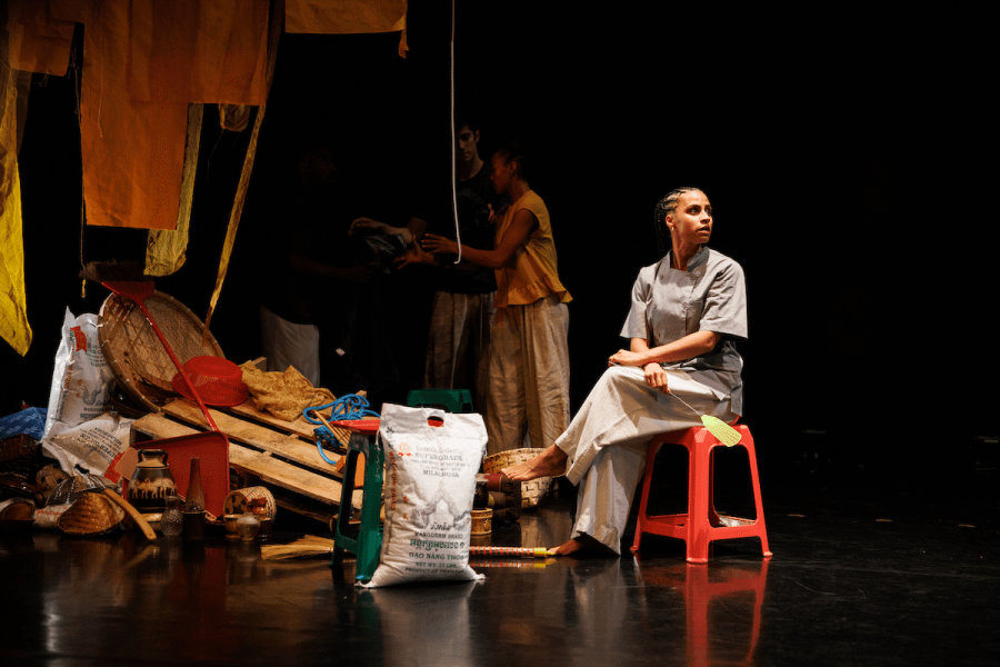 Three people are gathered by a pile of junk on a dark stage. One of them is seated on a red stool and looking off into the distance while the others inspect the junk.