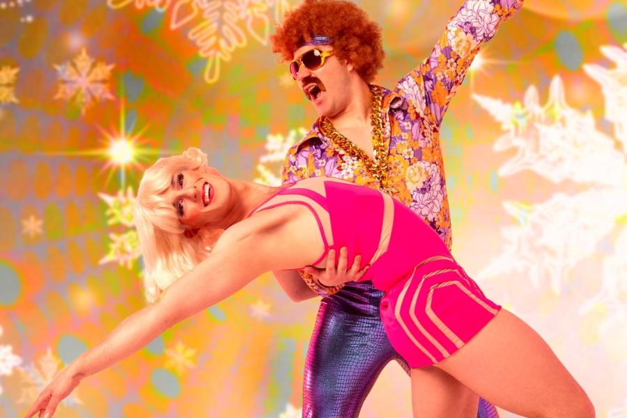 Two colorfully dressed performers dance together in front of a background of gold and snowflakes.