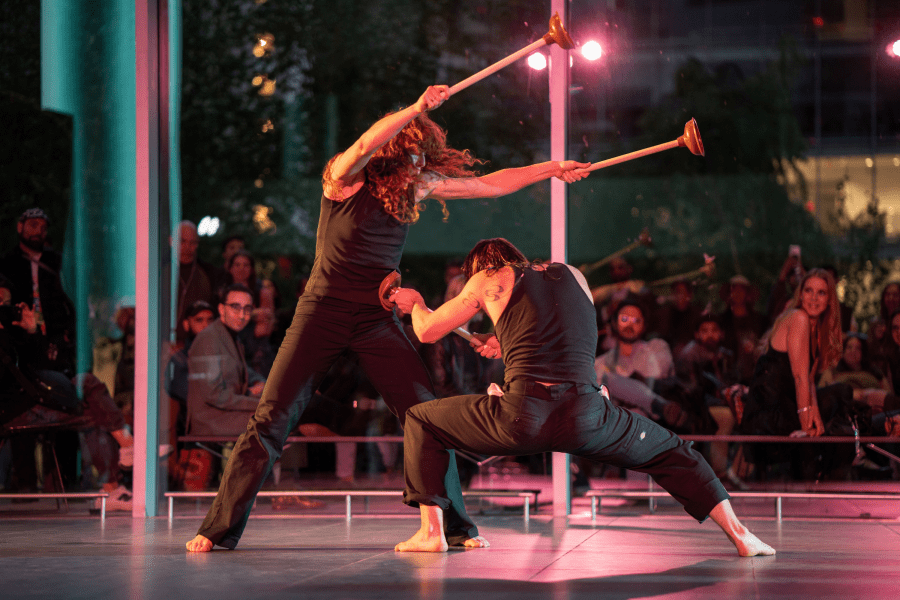One person holds out two toilet plungers in the air while a crouching person thrusts one against their stomach.