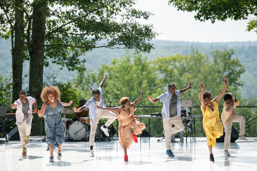 At an outdoor stage, seven Black dancers perform in a line.