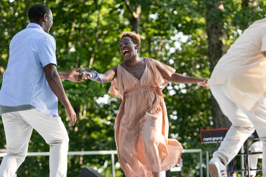 A Black female dancer holds her arm out to a Black male dancer. She has short, blonde-ish hair and wears a pink flowly jumpsuit.