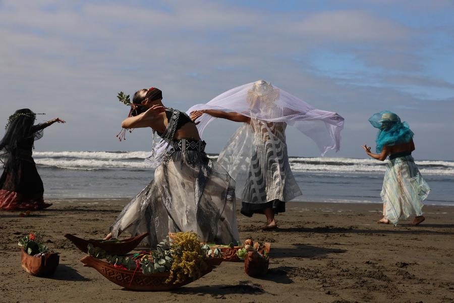 On a beach, folks wear masks and veils. Some spread their arms. One lays on the ground. 