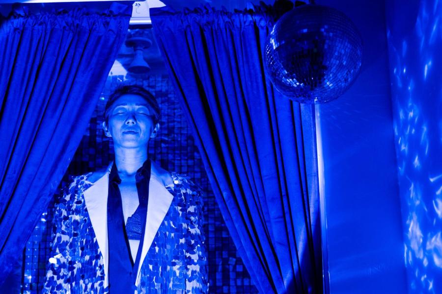 A person in a fringed jacket stands in front of a curtain in a room that is flooded with blue light.