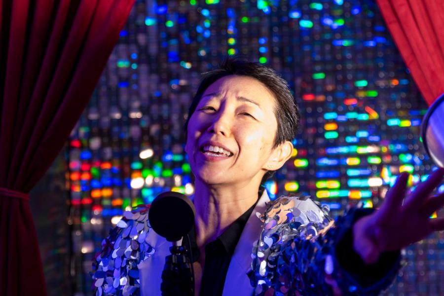 A person in a fringed jacket stands in front of a curtain and stretches out a hand.