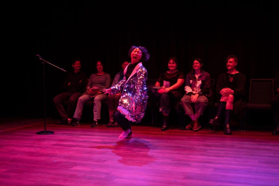 A person in a long fringed jacket yells on a stage as a seated audience watches.