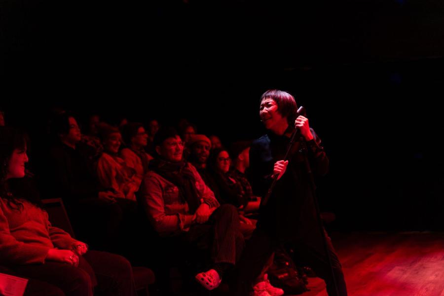 A person holding a short cane is observed by a seated audience.
