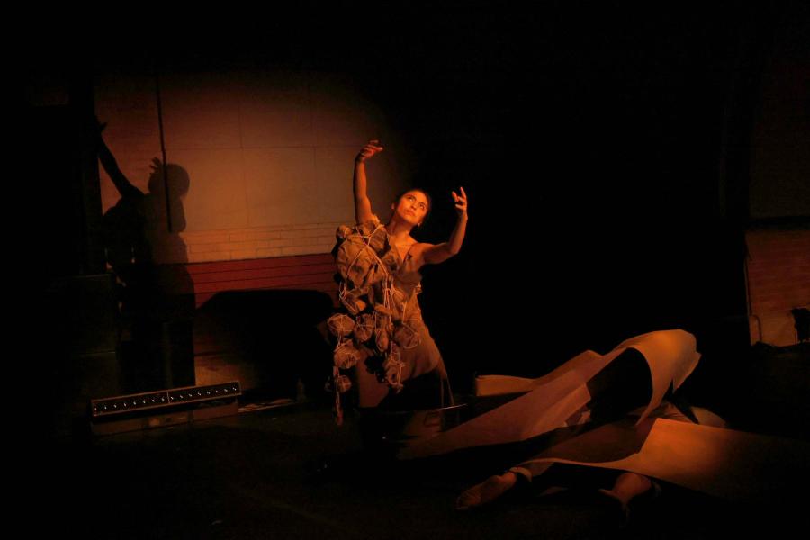 A kneeling performer lit by a spotlight raises their arms.