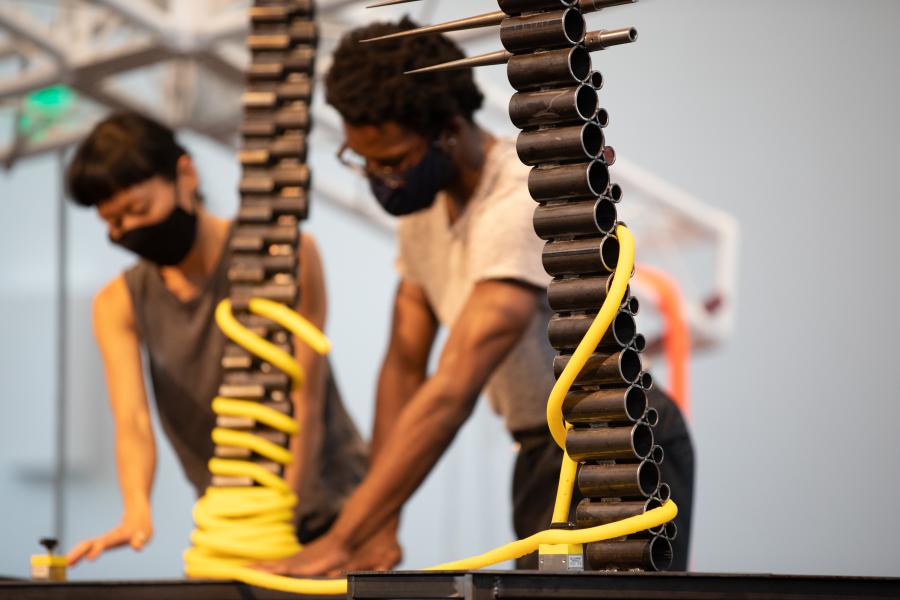 Two masked folks touch buttons, in the background, and, in the foreground, metal cylinders and yellow rope are intertwined.