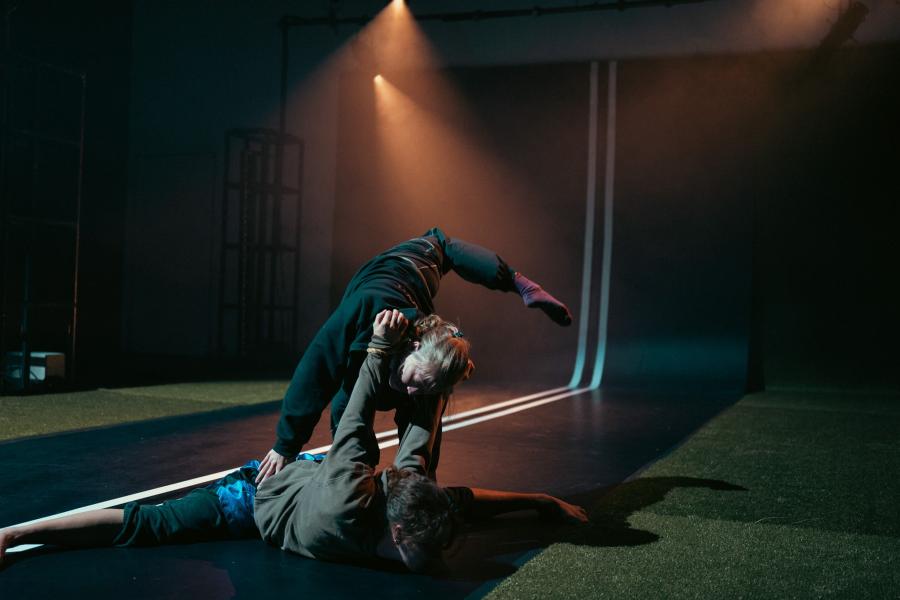 On a road that continues up the wall, a dancer "punches" another dancer on the ground, with stage lighting.