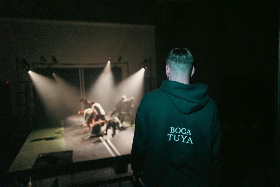 A dancer watches a group perform in stage lights and on a road that continues up the wall.