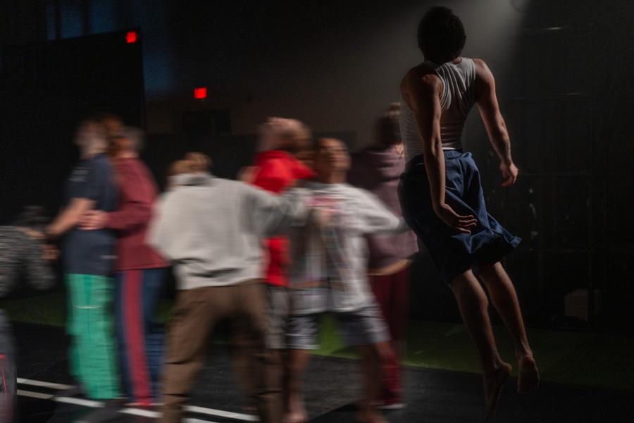 A group of dancers are motion-blurred, a dancer, in focus, leaps int the air.