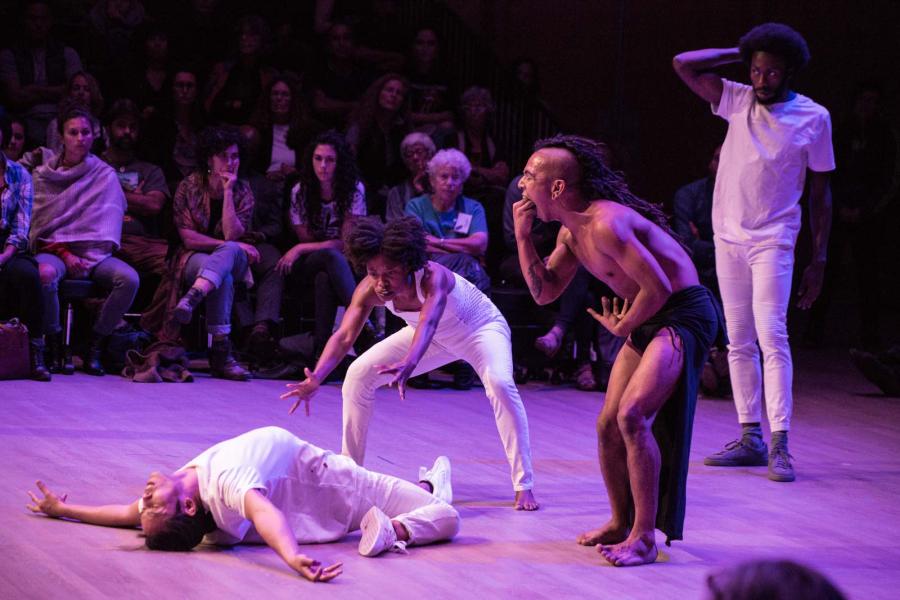 A man dressed in Native American garb pretends to vomit on two women who are dancing in white and look to be exorcising a demon from one of the women