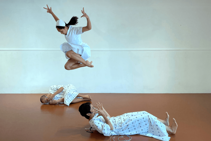 In a sterile space with off-white walls and a brown floor, two folks dressed in hospital gowns lay on the ground. A woman in a nurse's outfit leaps over one of them.