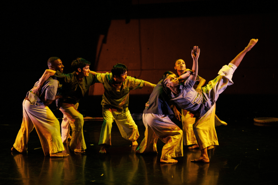 A group of six performers are on a stage with their arms slung over each others shoulders. The one on the far right raises a leg and arm towards the sky.