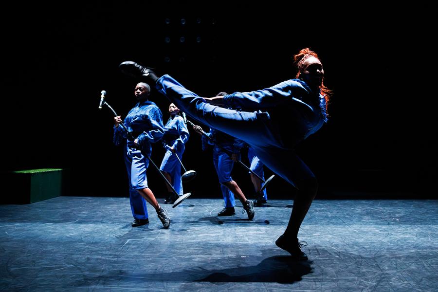 On a dark, overhead-lit stage, dancers, in casual wear, throw their legs up in different directions.
