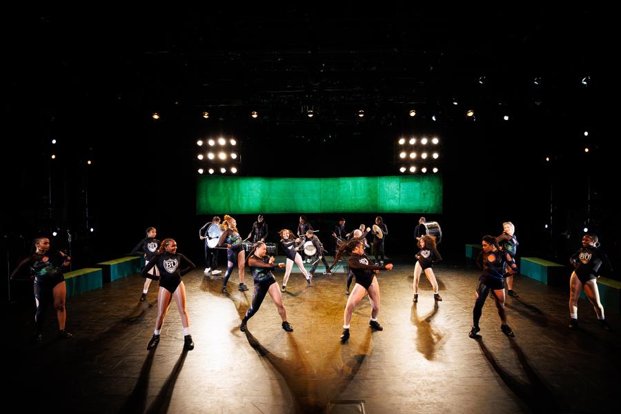 On a stage, a group of dancers in different stylized leotards perform with a green band of light behind them.