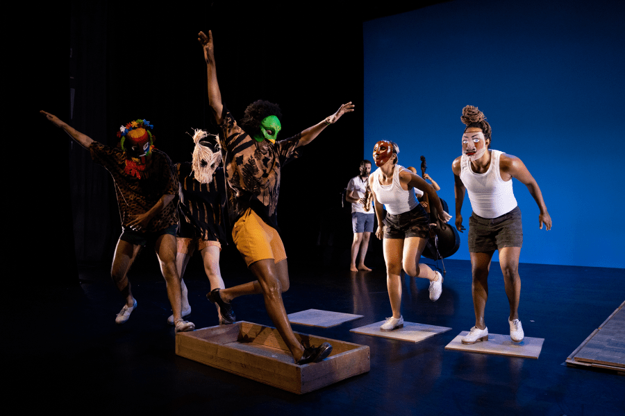 Masked performers leap around on a stage with a blue backdrop.