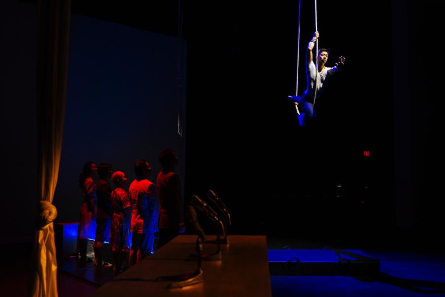 A performer swings from a rope, while other performers stand below on a stage in red light.