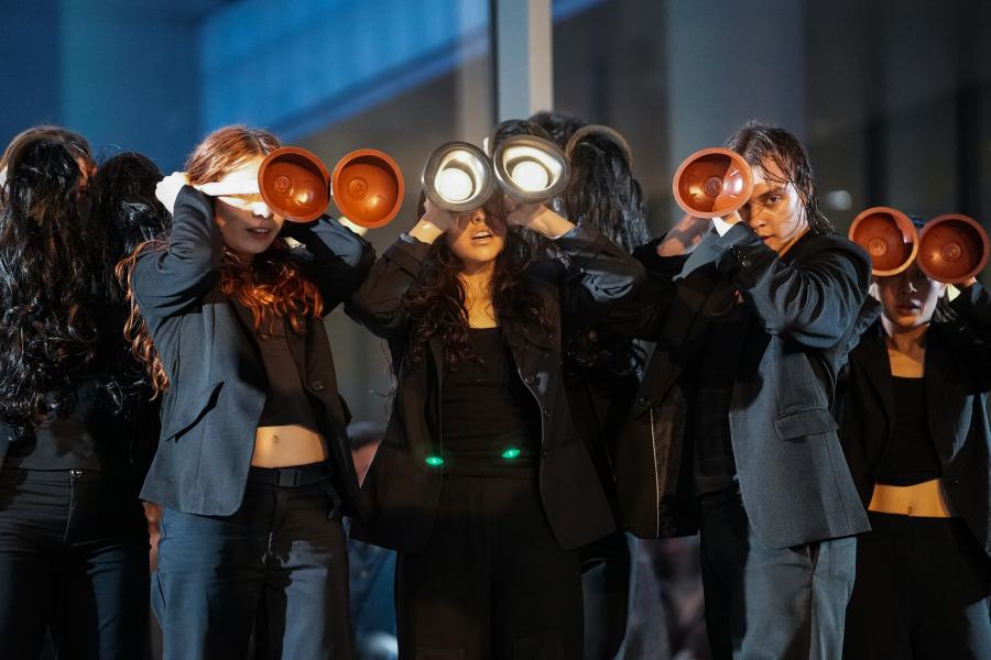 Three people dressed in black hold toilet plungers in front of their faces.