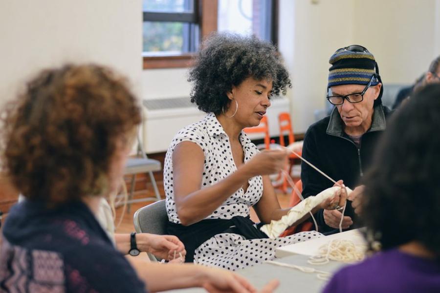 An older black woman knits in a crowd of folks.