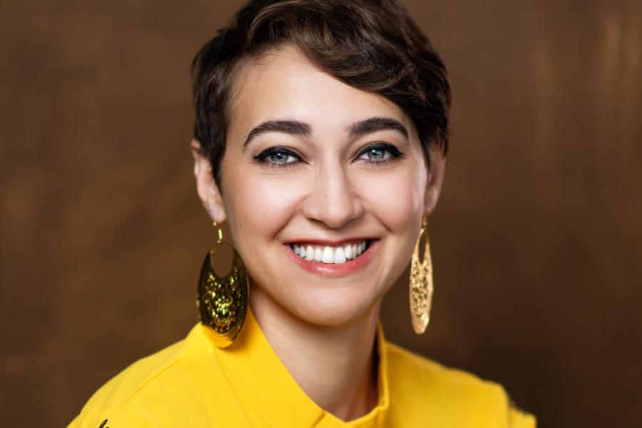 In front of a brown backdrop, a woman in gold earrings and a yellow sweater smiles.