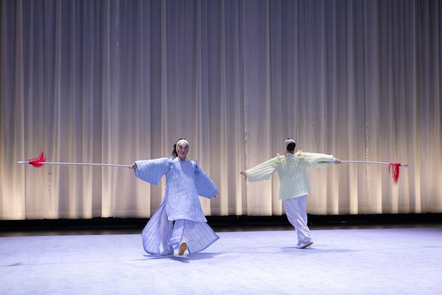 On a stage with a white curtain behind them, two performers hold white sticks with red strings at the end and face each other while walking in opposite directions, but possibly in a circle.