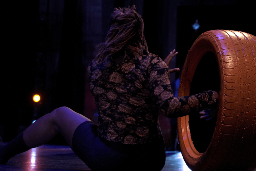 A photo taken from behind of a performer sitting on a stage with their right hand sticking into an upright orange tire.