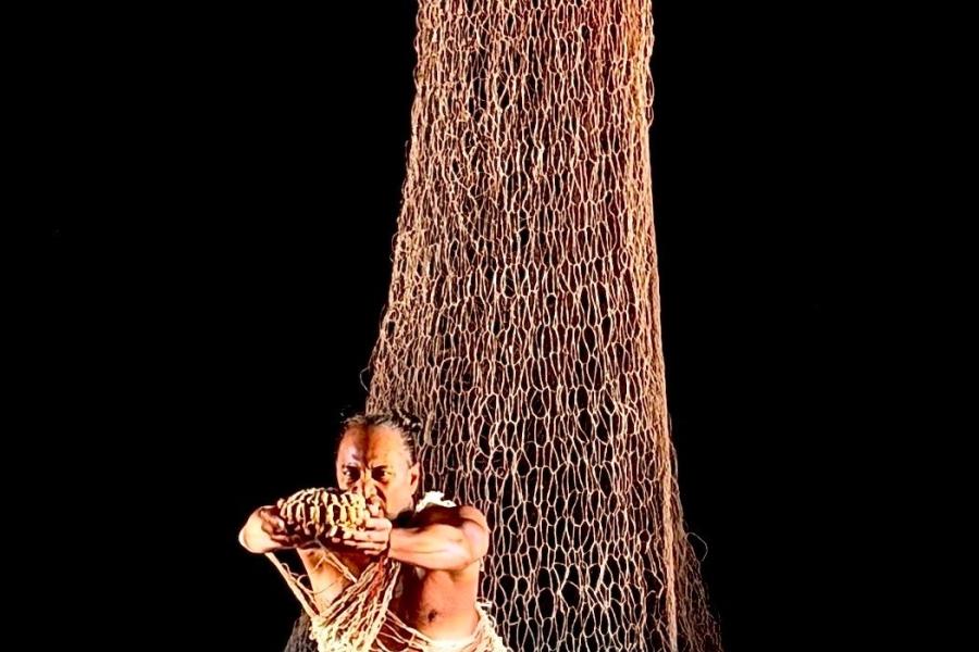 On a dark stage, a person stands in front of a net that drapes from the ceiling.