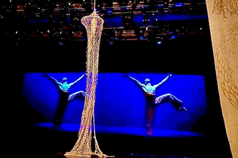 In the foreground a net descends from the ceiling. Further back a screen shows two performers stretching out their arms while raising a leg.