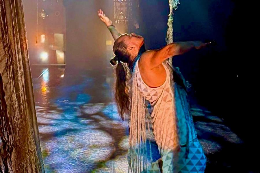 A performer holds onto a rope which descends from the ceiling while stretching out an arm.