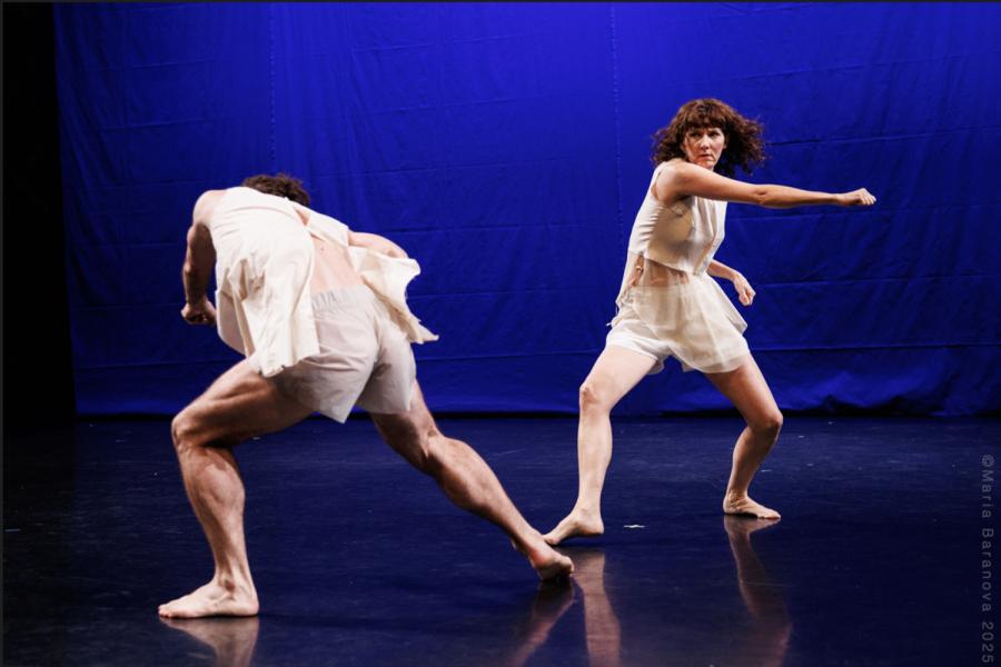 On a stage, with a blue background, two performers hold their feet near each other while one sings and the other ducks. The person who ducks has their back to us and the person who swings faces us.