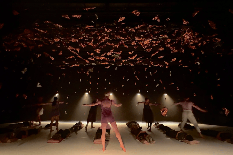 Half a dozen people stand on a stage while a dozen lie down on it. Scraps of foil dangle from the ceiling overhead.