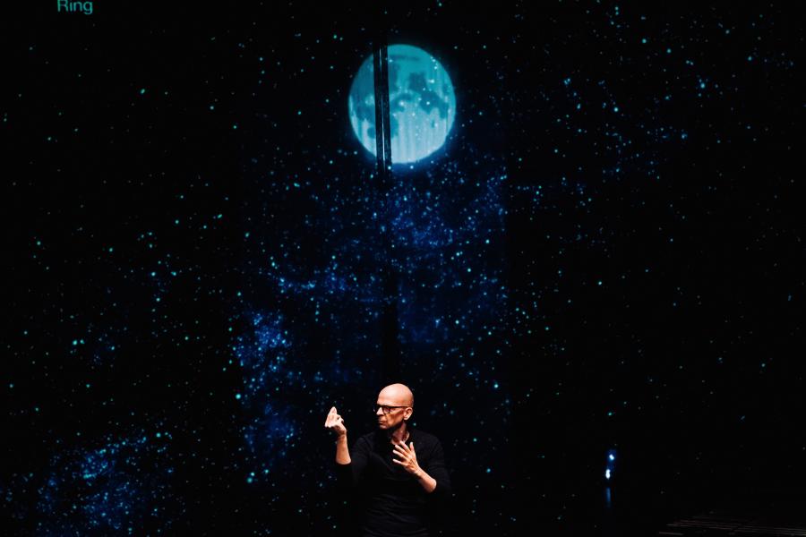 A white man signs in American Sign Lanuguage, before a projection of a blue moon.