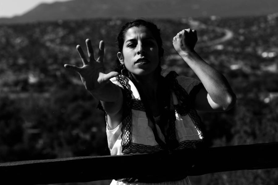 In black and white, a woman, with shoulder length dark hair, dances in the desert. Mountains behind her.